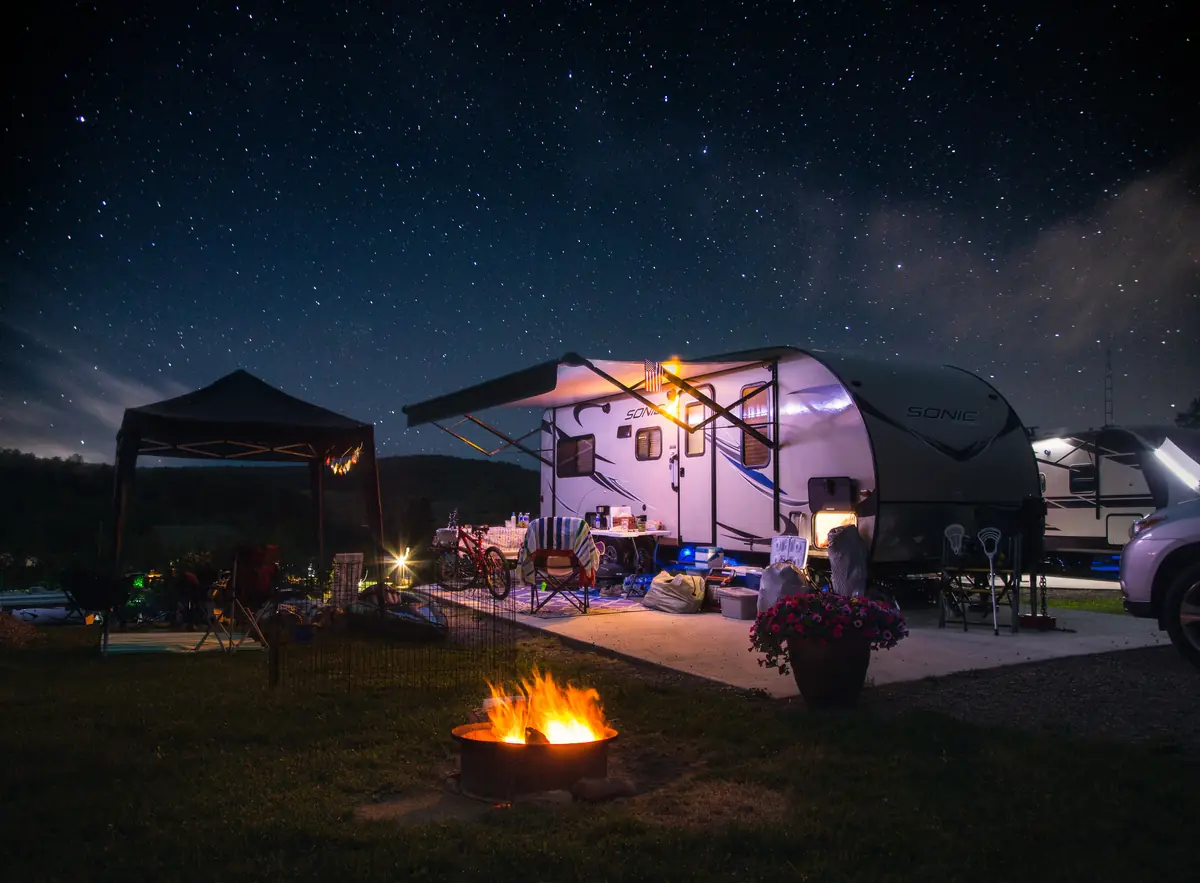 Travel trailer at a campsite under a starry night sky, with a campfire burning in a fire pit